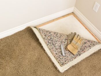Construction Gloves And Utility Knife On Pulled Back Carpet And Pad In Room.